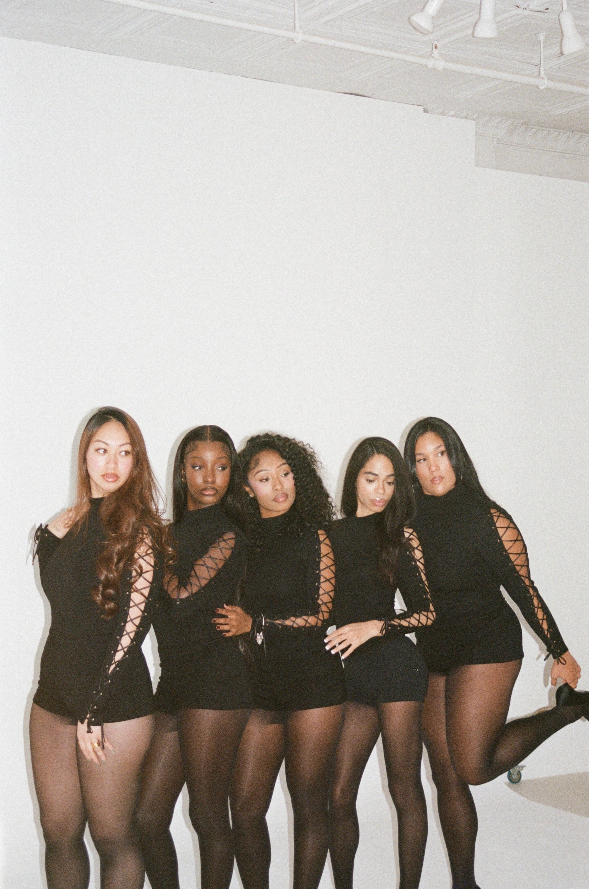Five women in black outfits with lace details posing against a white wall.